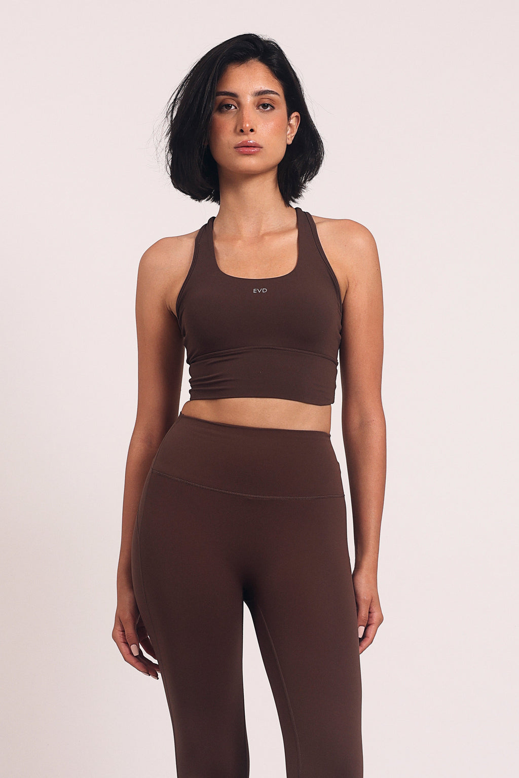 A woman modeling a matching dark brown activewear set consisting of a cropped tank top and high-waisted flared leggings, standing against a plain light background.