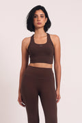 A woman modeling a matching dark brown activewear set consisting of a cropped tank top and high-waisted flared leggings, standing against a plain light background.