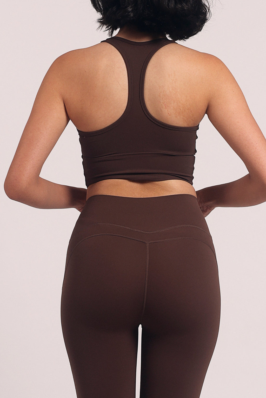 A woman shown from the back wearing a matching dark brown activewear set consisting of a sports bra and high-waisted flared leggings, standing against a solid light beige studio background.